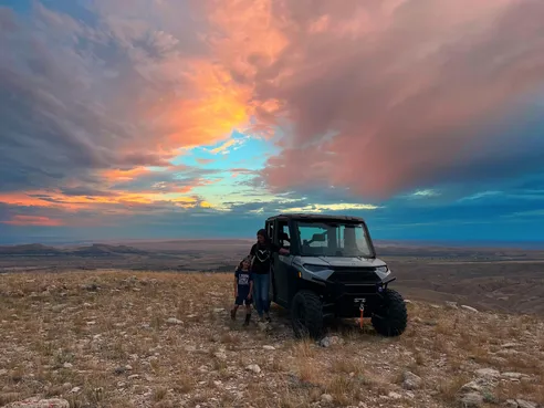 Emily and Case beside the side by side on a hilltop at sunset, the whole sky orange and pink over the basin below