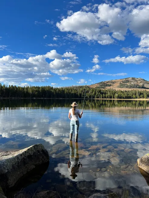 Emily in a wide brimmed hat and waders standing in the shallows of a mirror still mountain lake, casting a fly rod