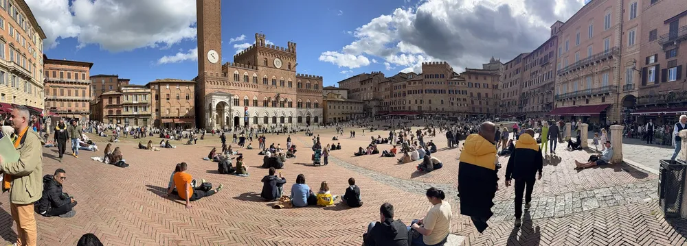 The Piazza del Campo in Siena, people sitting all over the sloping herringbone brick with the Torre del Mangia above the town hall