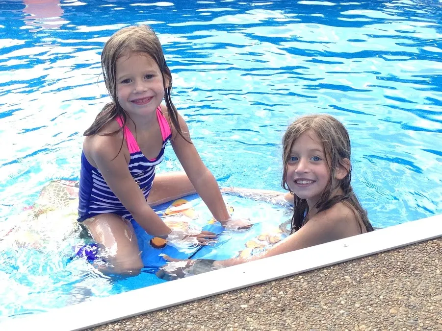 The two sisters in the pool at the edge, wet hair and big grins, one leaning on a float
