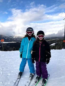 The two sisters side by side on their skis in helmets and goggles, cloud over the mountains behind