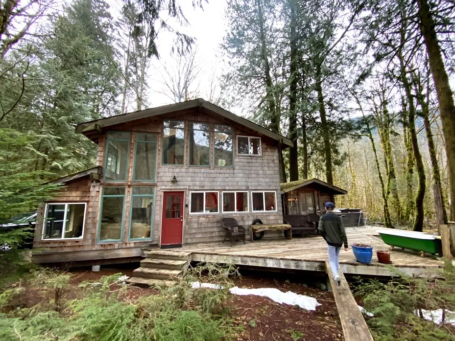 The Squamish cabin among tall firs, shingled walls and a red door, with a wooden deck across the front