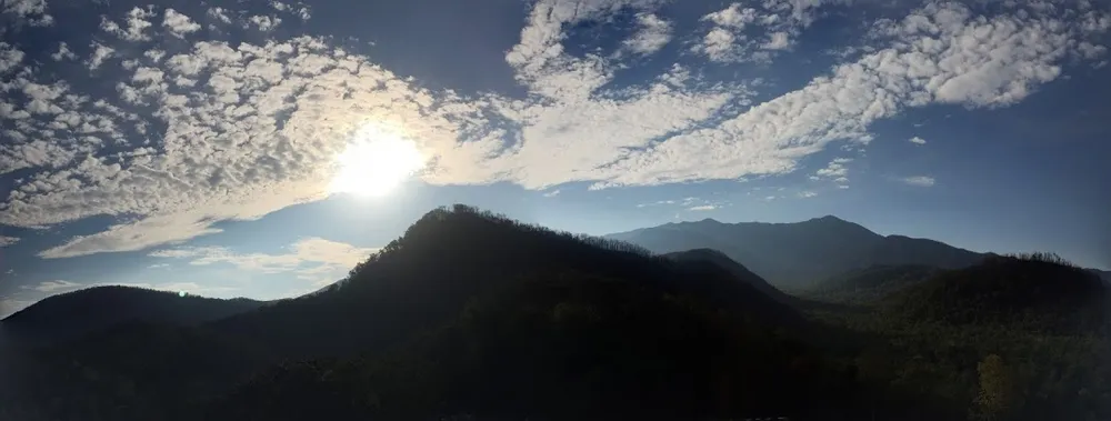 Sun breaking over a dark ridge of forested mountains under a wide sky of scattered cloud