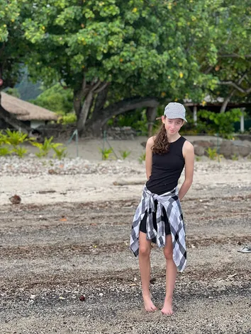Evie standing barefoot on the sand in a cap and a checked shirt tied at her waist