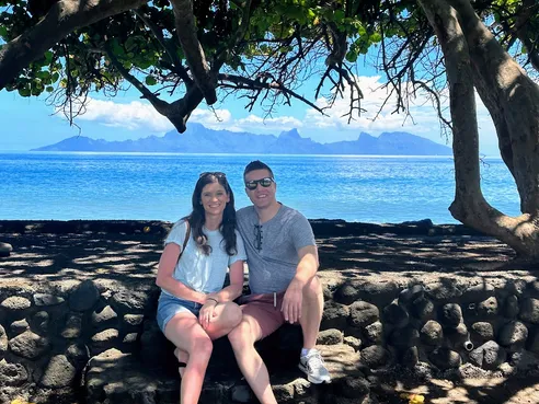 Emily and Josh sitting on a low stone wall in the shade, the blue lagoon and the peaks of Moorea behind