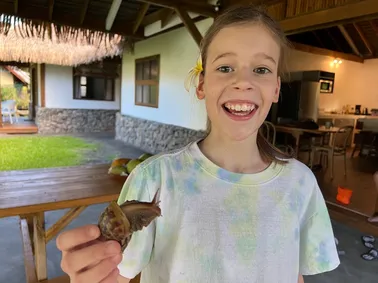Mia with a frangipani behind her ear, grinning and holding up a large snail