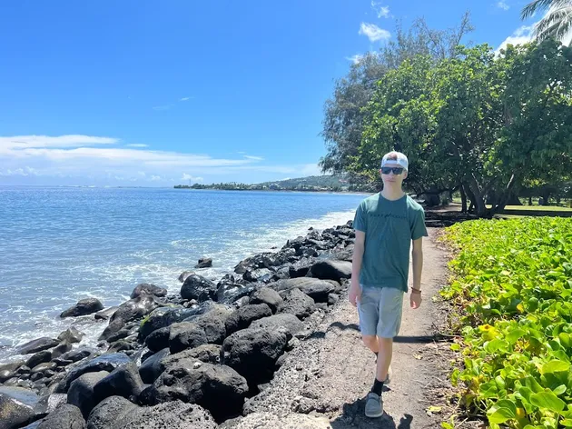 Preston walking a path between black volcanic boulders and green shore plants, the lagoon to his left