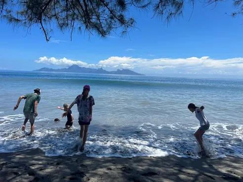Four of them playing in the surf on a black sand beach, Moorea on the horizon under white cloud