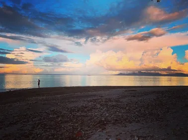 A lone figure at the water's edge on a dark shore, towering cloud lit pink and gold over the flat lagoon