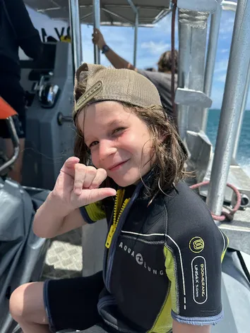 Case in a wetsuit and backwards cap on the dive boat, making a shaka sign at the camera