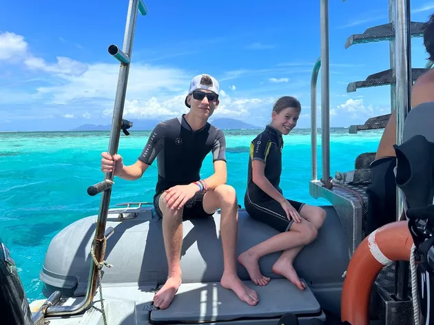 Preston and Mia in wetsuits sitting on the back of the dive boat, brilliant turquoise reef water behind them