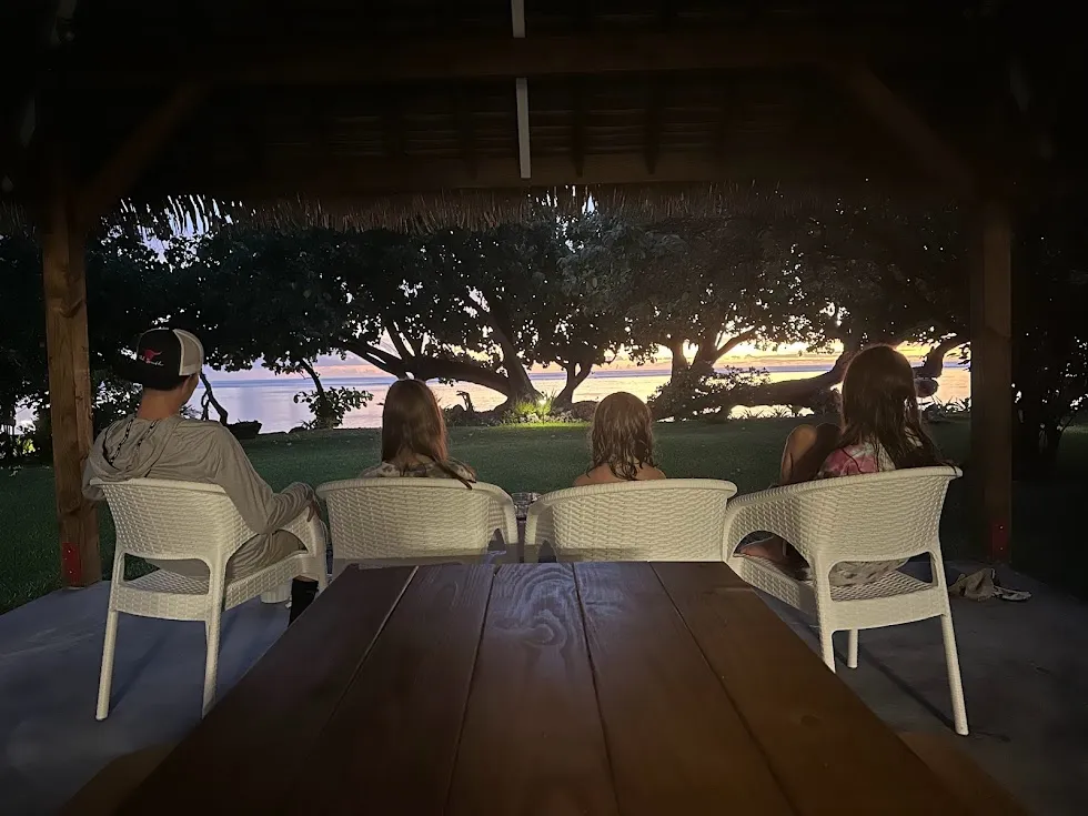 The four children in wicker chairs under the thatched shelter, all facing out to the water at dusk