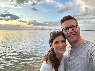 Emily and Josh close together at the water's edge, the lagoon and Moorea behind them at dusk