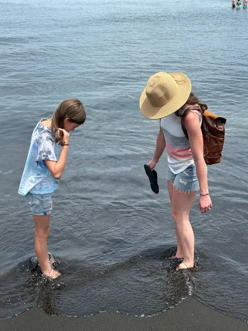 Emily in a wide straw hat and Mia standing ankle deep at the water's edge, both looking down at their feet in the black sand