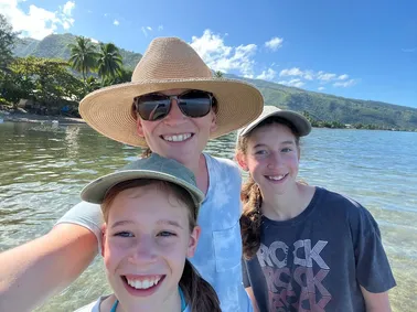 Emily in a wide straw hat with Mia and Evie, all three in the shallows with the green ridge behind