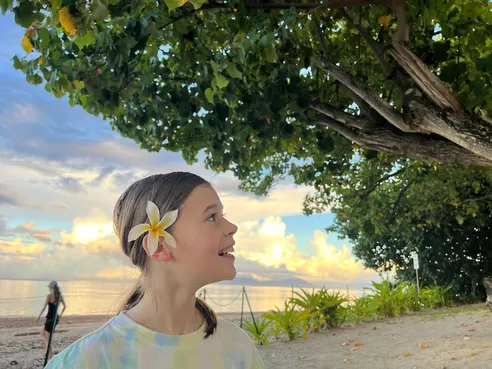 Mia with a frangipani behind her ear looking up and laughing, the beach and evening cloud behind her