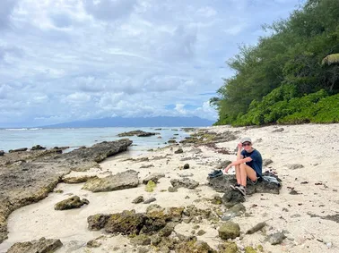 Evie sitting on a rock on a white coral beach, the reef and a distant island across the water