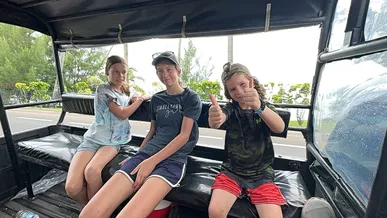 Three of the children on the bench seats in the back of an open safari truck, the youngest giving two thumbs up