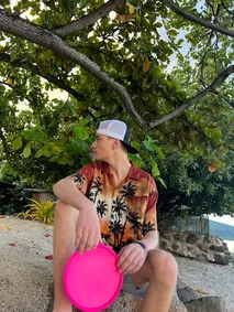 Preston in a palm-print shirt sitting under a spreading tree with a pink frisbee on his knee