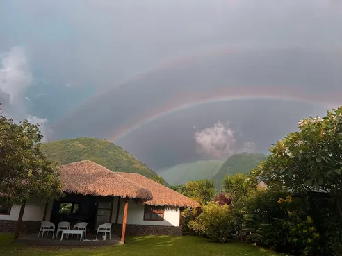 A double rainbow over the green ridge behind the thatched villa, chairs set out on the terrace below