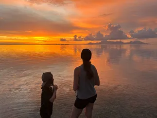 Emily and the youngest standing at the water's edge in silhouette, watching an orange sunset over Moorea