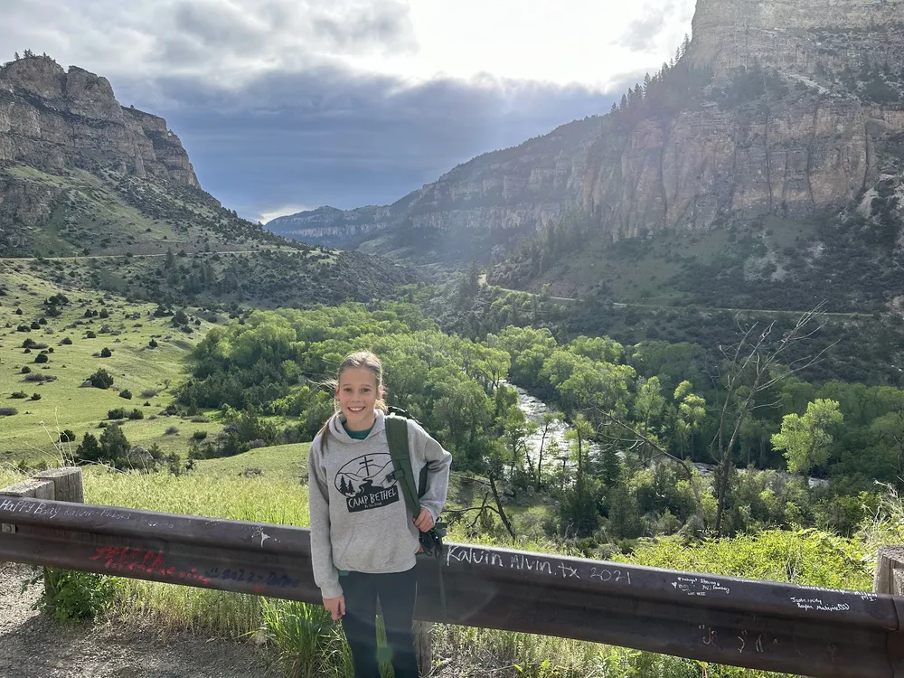 Mia with a daypack at a canyon overlook, leaning on a guardrail scratched with names, the creek and cliffs of Ten Sleep behind her
