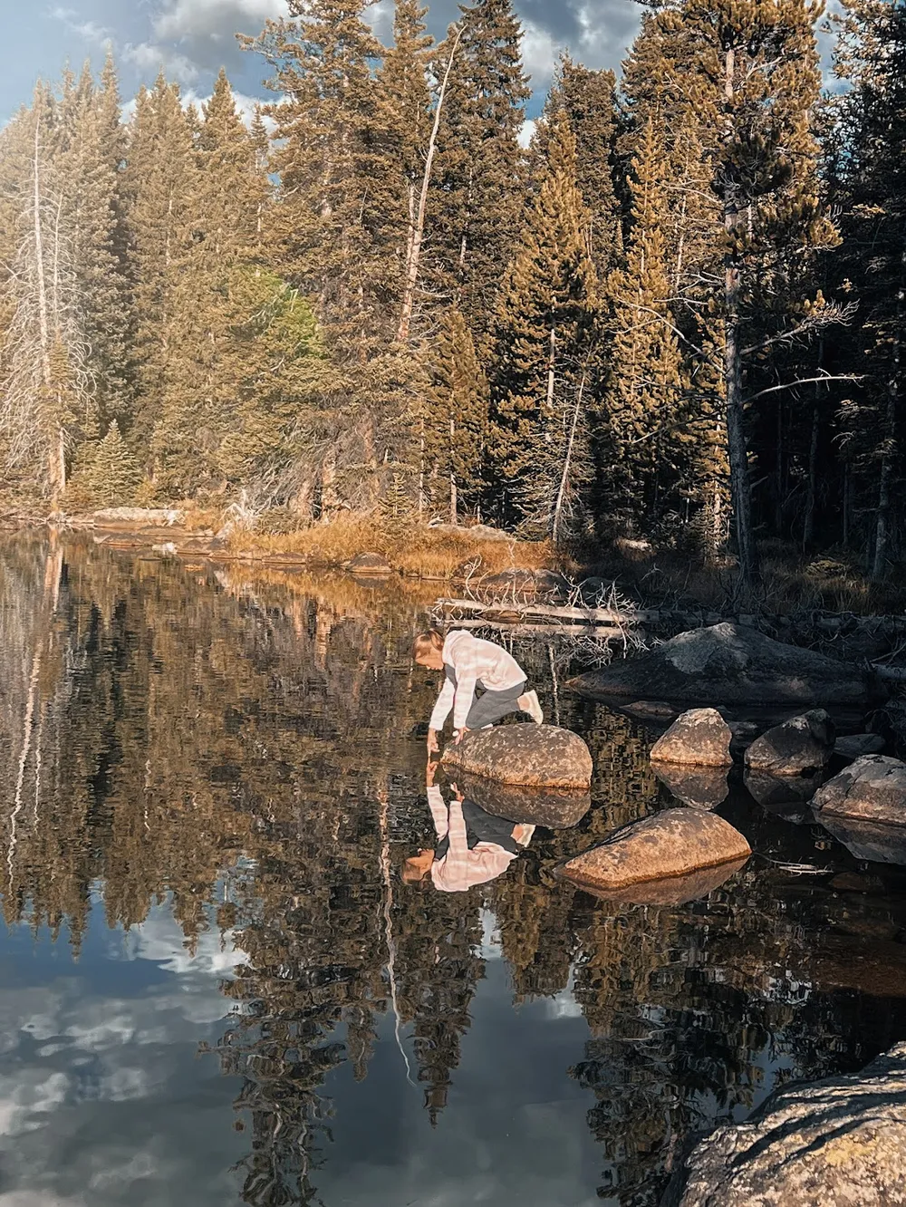 Mia bending to the edge of a still mountain lake, her reflection and the pines mirrored exactly in the water