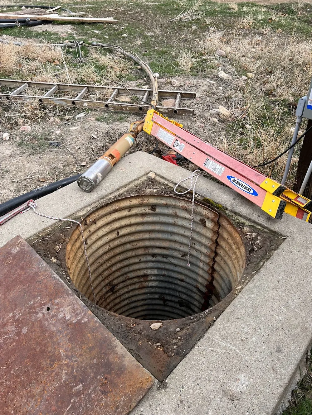 An open well shaft in a concrete pad with the pump pulled out and laid on top, a ladder braced across it