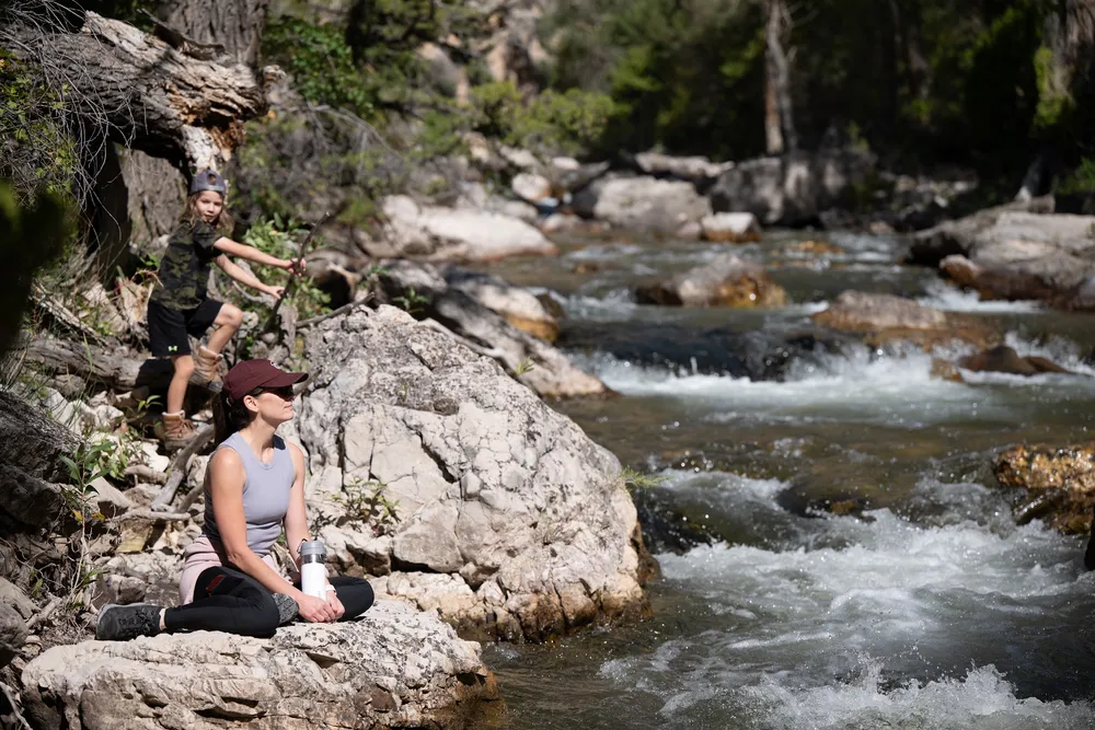 Emily sitting cross legged on a rock beside Shell Creek with a water bottle, Case climbing the bank behind her, white water running past