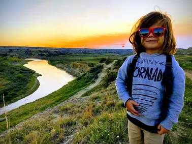 The youngest in red sunglasses and a Tiny Tornado sweatshirt on a bluff above a winding river, badlands and a gold sunset behind