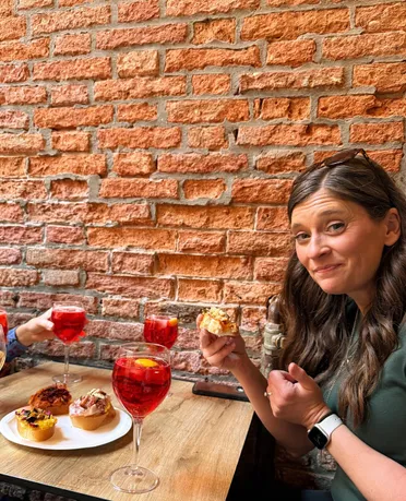 Emily at a bar table against bare brick holding up a cicchetto, a plate of them and glasses of red spritz in front of her