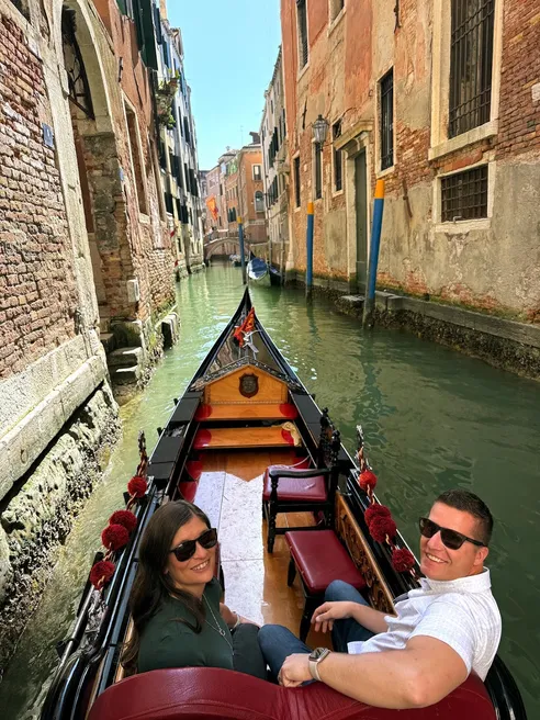 Emily and Josh turning round from the front seats of a gondola, a narrow green canal between old brick walls ahead
