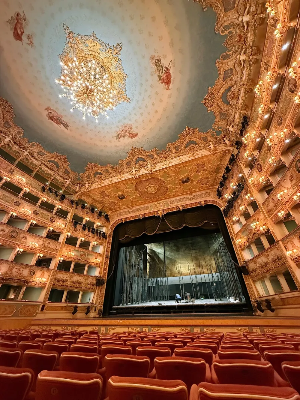 The gilded interior of La Fenice, tiers of boxes and a chandelier on the painted ceiling above rows of red seats