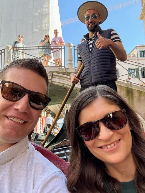 Emily and Josh in the gondola looking up at the camera, the gondolier in a straw hat giving a thumbs up behind them