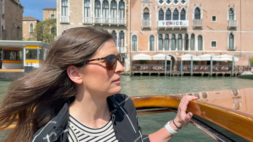 Emily in sunglasses on the back of a wooden water taxi, hair blown back, a Gothic palazzo on the Grand Canal behind her
