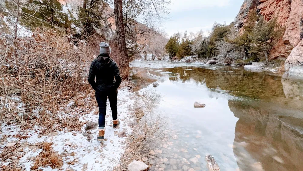 Emily in a winter coat and knit hat, seen from behind, walking a snowy bank beside a half frozen creek in a red rock canyon