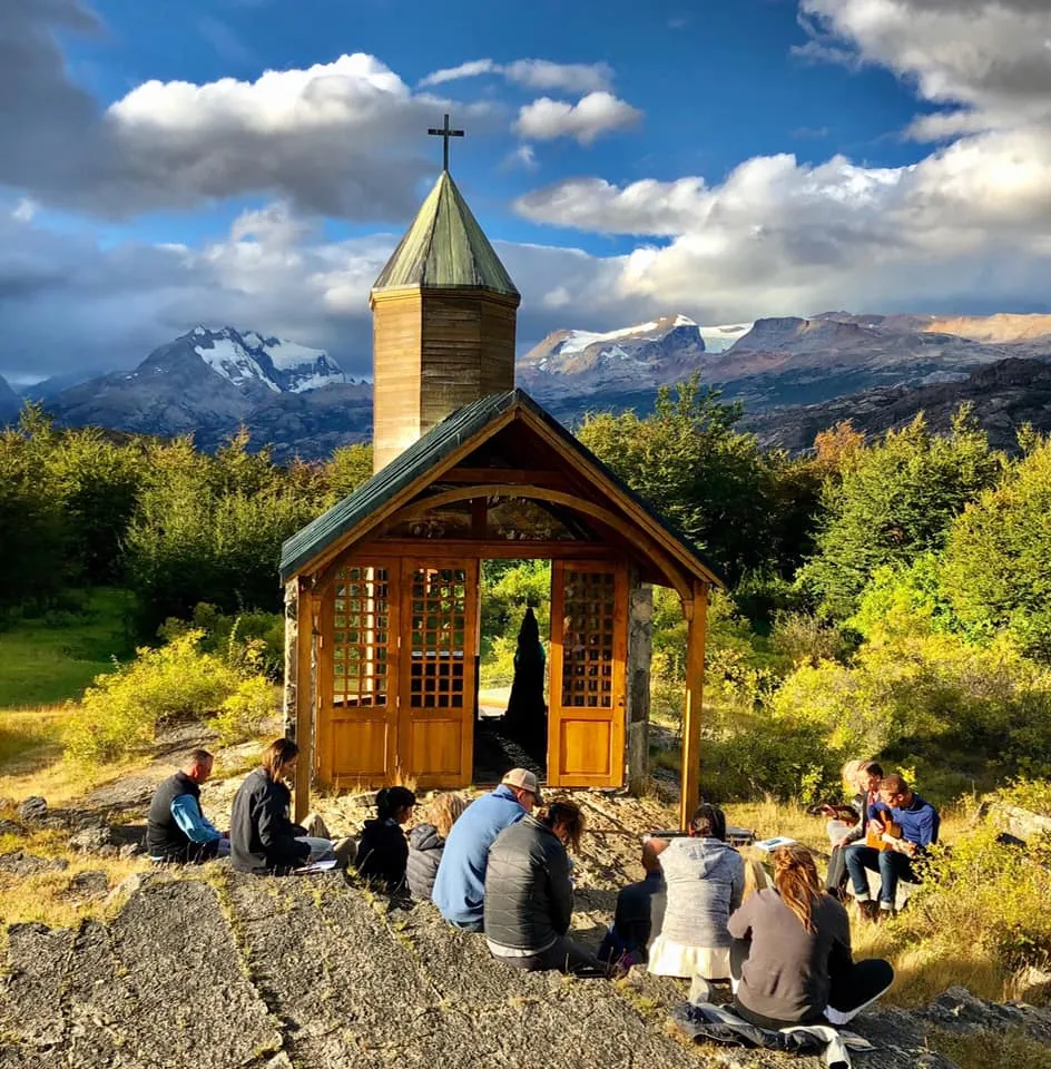 A group sitting on driftwood logs on a beach for a worship service, the glacier and mountain across the water
