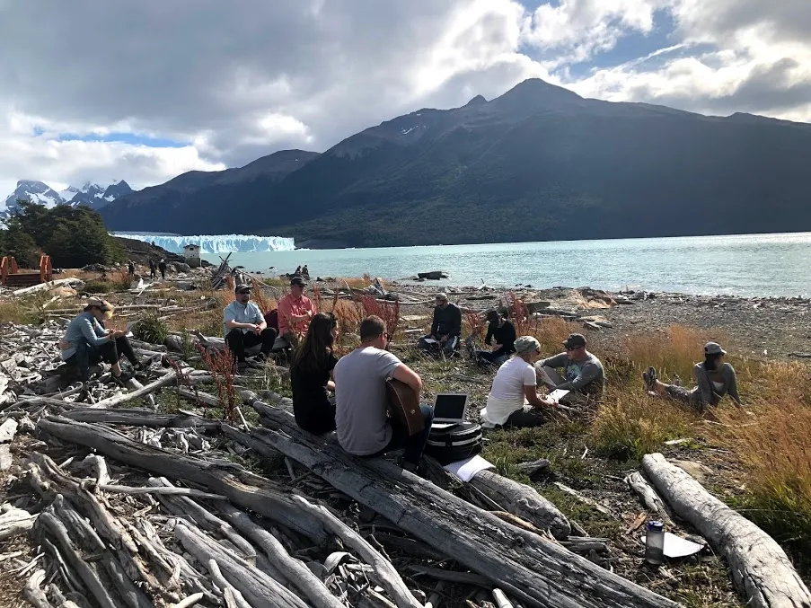 A gathering of the travel group seated on the beach with the glacier lake and snow peaks behind