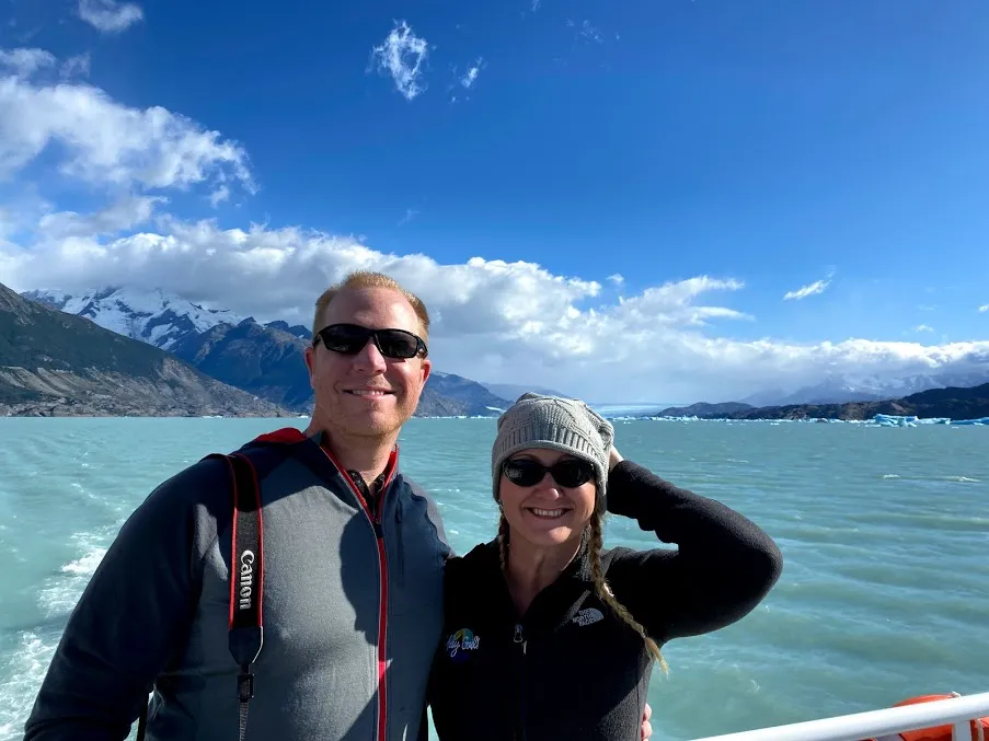 Chip and Heather in wetsuits by the pale blue glacial lake with snow-capped mountains rising behind