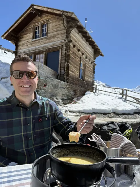 Josh dipping bread into a pot of cheese fondue on a terrace table, an old timber chalet and snow behind him