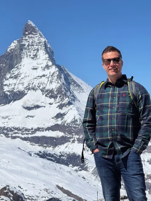 Josh in a checked shirt and daypack with the Matterhorn filling the sky behind him