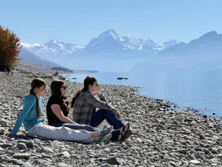Emily sitting on the stony shore of Lake Pukaki between Mia and Evie, all three looking out at Aoraki Mount Cook across the water