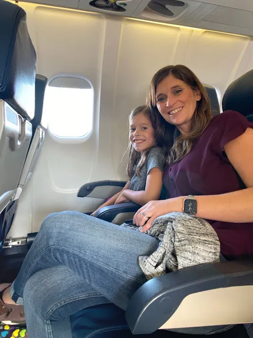 Emily and Mia buckled into their aeroplane seats, both smiling at the camera beside the window