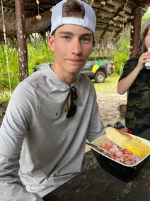 Preston at a picnic table under a palm shelter with a tray of poisson cru, bread and pineapple in front of him