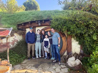 All six of them in the doorway of a hobbit hole at Hobbiton, grass roof above and a Baker Fresh Bread sign to one side